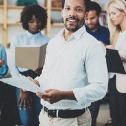 Teamwork concept in modern office.Young african businessman wearing white shirt holding papers at hands and standing front of the coworkers team.Vertical,blurred background